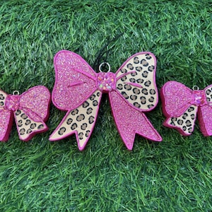 May include: Three pink glittery bow ornaments with a leopard print pattern. The bows are arranged on a green, grassy surface. The largest bow is in the center, with two smaller bows on either side.
