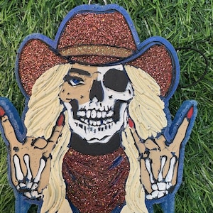 May include: A novelty ornament featuring a skull-cowboy design. The ornament has a red glitter cowboy hat, a red glitter bandana, and a blue border. The skull has blonde hair and is making a rock-and-roll hand gesture. The fingernails are painted red.