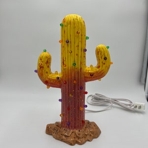 May include: A ceramic cactus with a yellow and red ombre paint finish and colorful lights. The cactus is on a brown base and has a cord for plugging in.
