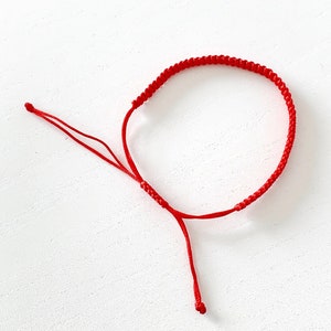 May include: A red string bracelet with a simple knot design. The bracelet is adjustable with a sliding knot.