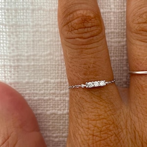 May include: A delicate silver ring with three small, clear gemstones in a row. The ring is attached to a fine chain, creating a minimalist design. The ring is worn on a finger, showcasing its subtle sparkle. The ring is a piece of jewellery.