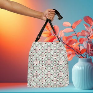 May include: A tote bag featuring a repeating geometric pattern of small red flowers, green leaves, and intricate white and tan square motifs. The bag has black adjustable straps with metal hardware, providing a functional design for carrying items.