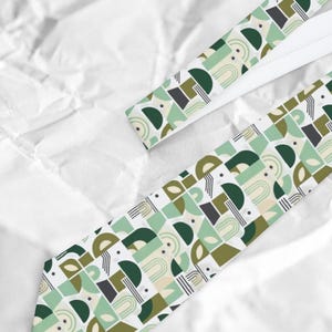 May include: A necktie featuring a repeating geometric pattern of semicircles, arches, and lines in shades of dark green, olive green, light mint green, and cream on a white background. The design includes small black dots and thin parallel lines.