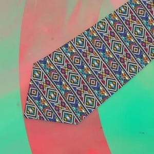 May include: A patterned necktie with a geometric design. The tie features a repeating pattern of colorful diamonds and rectangles in shades of blue, yellow, red, and green on a black background. The tie is angled across a background of pink and green.