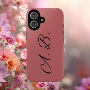 May include: A dusty rose mobile phone case featuring a black camera module with two lenses and a small flash. The back of the case is printed with the black cursive initials A. B. positioned vertically along the right side of the phone case.