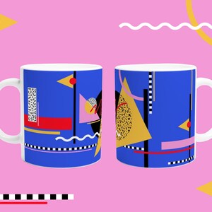 Op de afbeelding: Witte keramische mok met een levendig abstract ontwerp. Het ontwerp bevat geometrische vormen in blauw, geel, rood, zwart en roze tegen een witte achtergrond. De mok heeft een gebogen handvat en een glanzende afwerking. Het ontwerp herhaalt zich aan beide zijden.