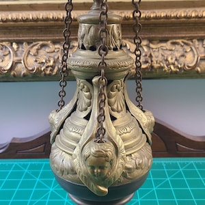 May include: A gold-toned metal incense burner with intricate details and cherub figures. The burner hangs from chains and has a black base.
