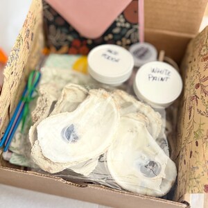 May include: A craft kit in a cardboard box containing several natural oyster shells, two small white plastic jars labeled White Paint and Mod Podge, a set of thin blue and green brushes, and a patterned fabric pouch with a pink top section.