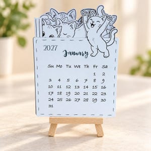 May include: A 2027 January desk calendar on a small wooden easel. The white card features a black and white illustration of multiple cats, with one cat standing on the right side. The calendar grid displays the days of the month clearly.