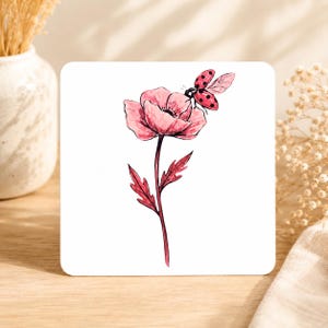 May include: Square white coaster featuring a watercolor illustration of a pink poppy flower and a ladybug. The flower has a dark center and is set against a white background. The ladybug has red wings with black spots.