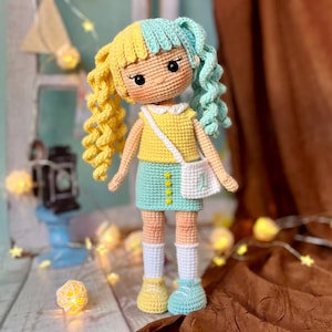 May include: A crocheted doll with yellow and blue hair, wearing a yellow shirt, a green skirt, and white socks. The doll is holding a white bag with a zodiac symbol.