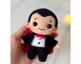 Op de afbeelding: Gehaakte vampierpop met een zwart pak, rode vlinderdas en cape. De pop heeft een lachend gezicht, zwart haar en grote zwarte ogen. De tekst "ENGLISH CROCHET PATTERN" staat bovenaan.