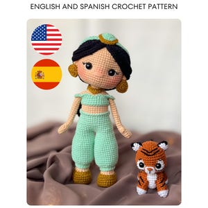 Pode incluir: Uma boneca de crochê com cabelo preto, top e calças verde menta, acessórios dourados e um pequeno tigre laranja e branco. O texto diz ENGLISH AND SPANISH CROCHET PATTERN, acompanhado pelas bandeiras dos Estados Unidos e da Espanha.