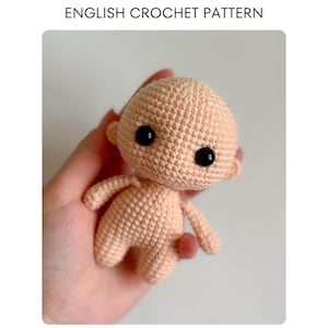 May include: A peach-colored crocheted doll with black button eyes and small ears. The doll has a rounded head, small arms, and legs. The text "ENGLISH CROCHET PATTERN" is at the top of the image.