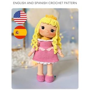 Può includere: Una bambola all'uncinetto con lunghi capelli gialli ricci, una piccola corona gialla e un vestito rosa con bordo bianco. La bambola ha occhi neri e guance rosate. Il testo in alto recita ENGLISH AND SPANISH CROCHET PATTERN, con le bandiere USA e Spagna.