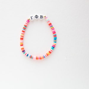 May include: A colorful beaded bracelet with the letters "ΓΦB" in black on white beads. The bracelet features a variety of bright, flat, disc-shaped beads in shades of pink, red, orange, yellow, blue, and green. The bracelet is on a white background.