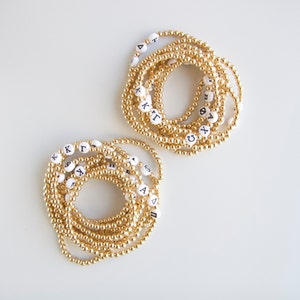 May include: Two sets of gold beaded bracelets with white beads that have Greek letters printed on them. The bracelets are stacked on top of each other.