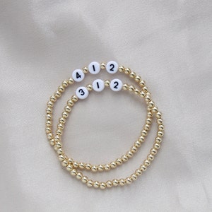 May include: Two gold beaded bracelets with white beads that have black numbers on them. The numbers on the bracelets are 1, 2, 3, and 4.