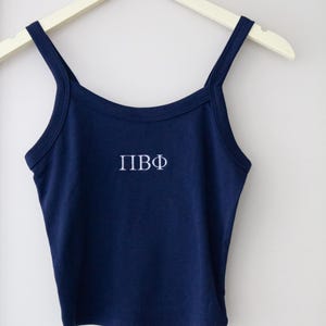 May include: A navy blue crop top with thin straps and the embroidered letters "ПВФ" in white. The top is hanging on a white hanger against a plain white background. The style is casual and simple.