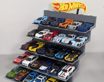 May include: A gray plastic Hot Wheels display stand with six shelves, each holding a row of colorful toy cars. The stand features the Hot Wheels logo at the top.