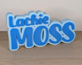 May include: A 3D-printed sign in light blue and dark blue, spelling out "Lachie MOSS". The letters are in a playful, rounded font, with a shadow effect. The sign is placed on a wooden surface.