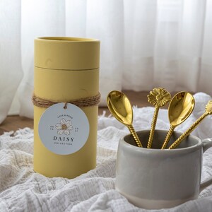 May include: A set of four gold-colored spoons with daisy-shaped tops in a gray mug. A yellow cylindrical container with a tag that reads "DAISY COLLECTION" is next to the mug. The spoons have twisted handles.