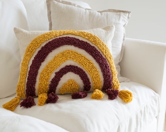 May include: A throw pillow with a rainbow design in shades of burgundy and yellow. The pillow has a textured, tufted surface and tassels on the edges.