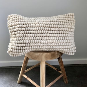 May include: A white, fluffy cushion with a textured, ball-like surface. The cushion is sitting on a wooden stool with three legs.