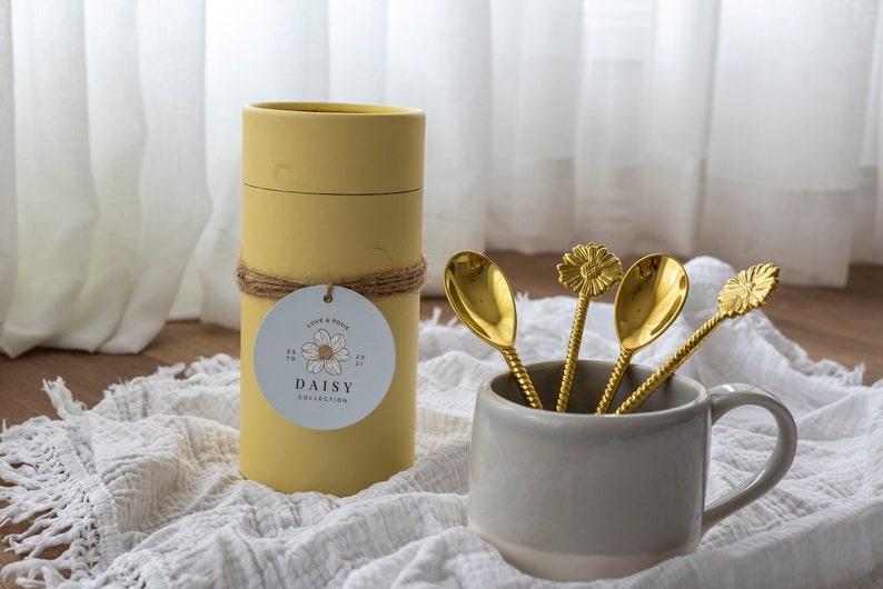 May include: A set of four gold-colored spoons with daisy-shaped tops in a gray mug. A yellow cylindrical container with a tag that reads "DAISY COLLECTION" is next to the mug. The spoons have twisted handles.
