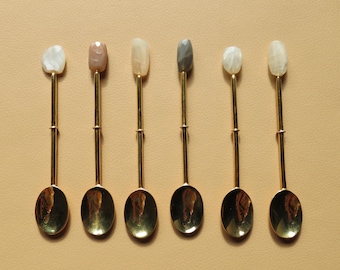 May include: Set of seven gold spoons with different colored gemstone handles. The handles are in shades of white, brown, gray, and pink.