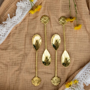 May include: Four gold-colored spoons with floral designs on the handles and spoon bowls. The spoons are arranged on a tan fabric with white lace trim and yellow flowers. The spoons have a twisted stem.