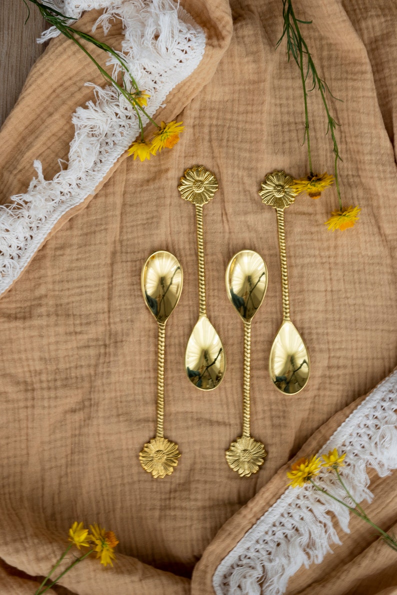 May include: Four gold-colored spoons with floral designs on the handles and spoon bowls. The spoons are arranged on a tan fabric with white lace trim and yellow flowers. The spoons have a twisted stem.