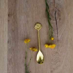 May include: A gold-colored spoon with a floral design on the handle, resting on a light brown wooden surface. Yellow flowers and green stems are arranged around the spoon. The spoon's bowl reflects the surrounding objects.
