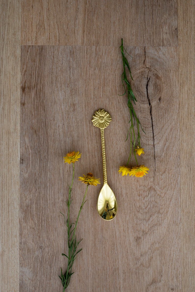 May include: A gold-colored spoon with a floral design on the handle, resting on a light brown wooden surface. Yellow flowers and green stems are arranged around the spoon. The spoon's bowl reflects the surrounding objects.