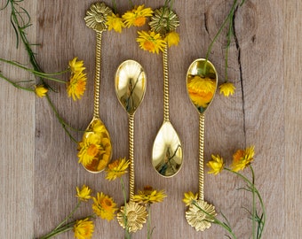 May include: Four gold-colored spoons with floral designs on the handles and bowls, arranged on a light brown wooden surface. Yellow flowers and green stems are artfully placed around the spoons, creating a decorative display. The spoons are approximately 6 inches long.