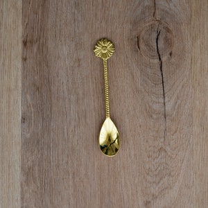 May include: A gold-colored spoon with a floral design on the handle and a teardrop-shaped bowl. The handle has a twisted rope design. The spoon is approximately 6 inches long and rests on a wooden surface.