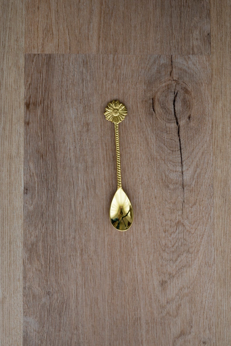 May include: A gold-colored spoon with a floral design on the handle and a teardrop-shaped bowl. The handle has a twisted rope design. The spoon is approximately 6 inches long and rests on a wooden surface.