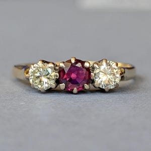 May include: A three-stone ring featuring a central round-cut deep red ruby flanked by two round-cut white diamonds. The stones are set in a polished yellow gold band with six-prong settings for each gemstone, creating a classic horizontal arrangement.