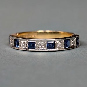 Puede incluir: Un anillo de banda de oro y plata con piedras preciosas azules cuadradas alternas y piedras preciosas claras más pequeñas. El anillo tiene un diseño clásico con un acabado pulido, adecuado para el uso diario u ocasiones especiales.