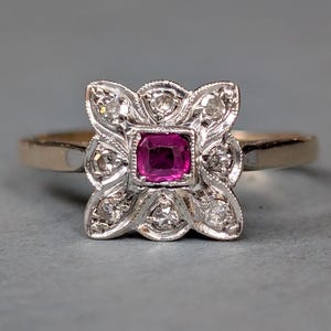 Puede incluir: Un anillo vintage con una piedra central cuadrada de rubí rosa intenso. El rubí está rodeado por un diseño floral de metal blanco, acentuado con pequeños diamantes. La banda es de color oro.