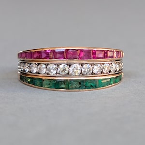 May include: Three gold rings stacked together. The top ring is lined with square-cut rubies, the middle ring with round diamonds, and the bottom ring with square-cut emeralds. The rings are set against a grey background.