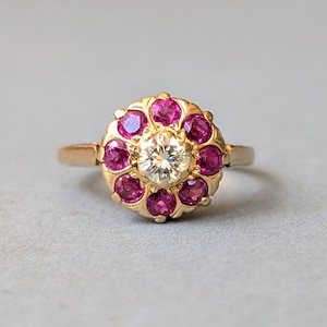 Puede incluir: Un anillo de oro con un diamante blanco redondo central rodeado por un halo de ocho rubíes rosas redondos. Las piedras están engastadas en una montura de oro festoneada que crea un diseño de racimo floral sobre una banda de oro lisa.