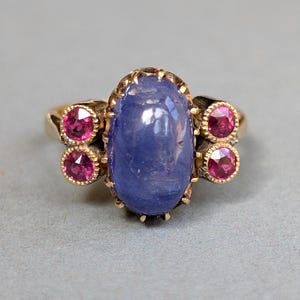 May include: A gold ring featuring a large, oval, blue gemstone as the centerpiece, flanked by two pairs of smaller, round, red gemstones. The ring has an ornate, vintage design with a detailed setting.