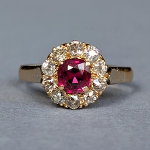 May include: A gold ring featuring a central cushion-cut deep pinkish-red gemstone surrounded by a halo of ten round-cut clear white diamonds. The band is smooth and polished gold, providing a classic setting for the vibrant center stone.