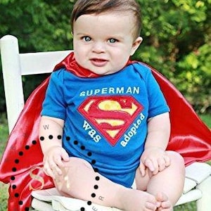 May include: A baby wearing a blue Superman onesie with a red cape, sitting on a white chair. The onesie has the Superman logo and the words "Superman was adopted". The baby is smiling, with a green background.