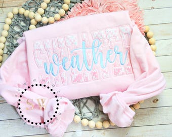 May include: A pink sweatshirt with the text "sweater weather" embroidered in blue and white on the front. The sweatshirt has a white and pink floral pattern.