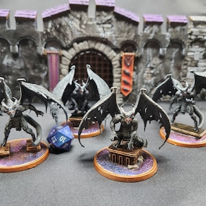 May include: Miniature fantasy figures of winged demons with horns and red eyes, posed on circular bases. The figures are dark gray with detailed wings, set against a backdrop of a castle wall and gate. A blue and white die is also present.