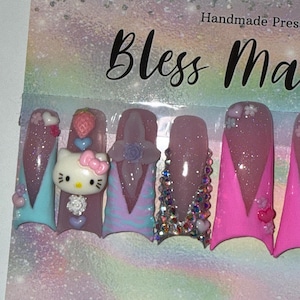 May include: A set of handmade press-on nails with a variety of designs. The nails feature a Hello Kitty design, glitter, rhinestones, and other decorative elements in shades of pink, purple, and blue. The text "Bless Ma" is visible.