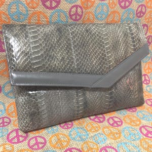 May include: A grey clutch bag with a snakeskin pattern and a grey flap closure. The bag is rectangular and has a sleek, modern design. The flap is a lighter shade of grey.