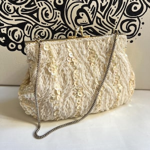 May include: Ivory beaded clutch bag with a silver chain strap. The vintage handbag features a clasp closure and is embellished with sequins. The bag has a rectangular shape and a gold-toned frame.